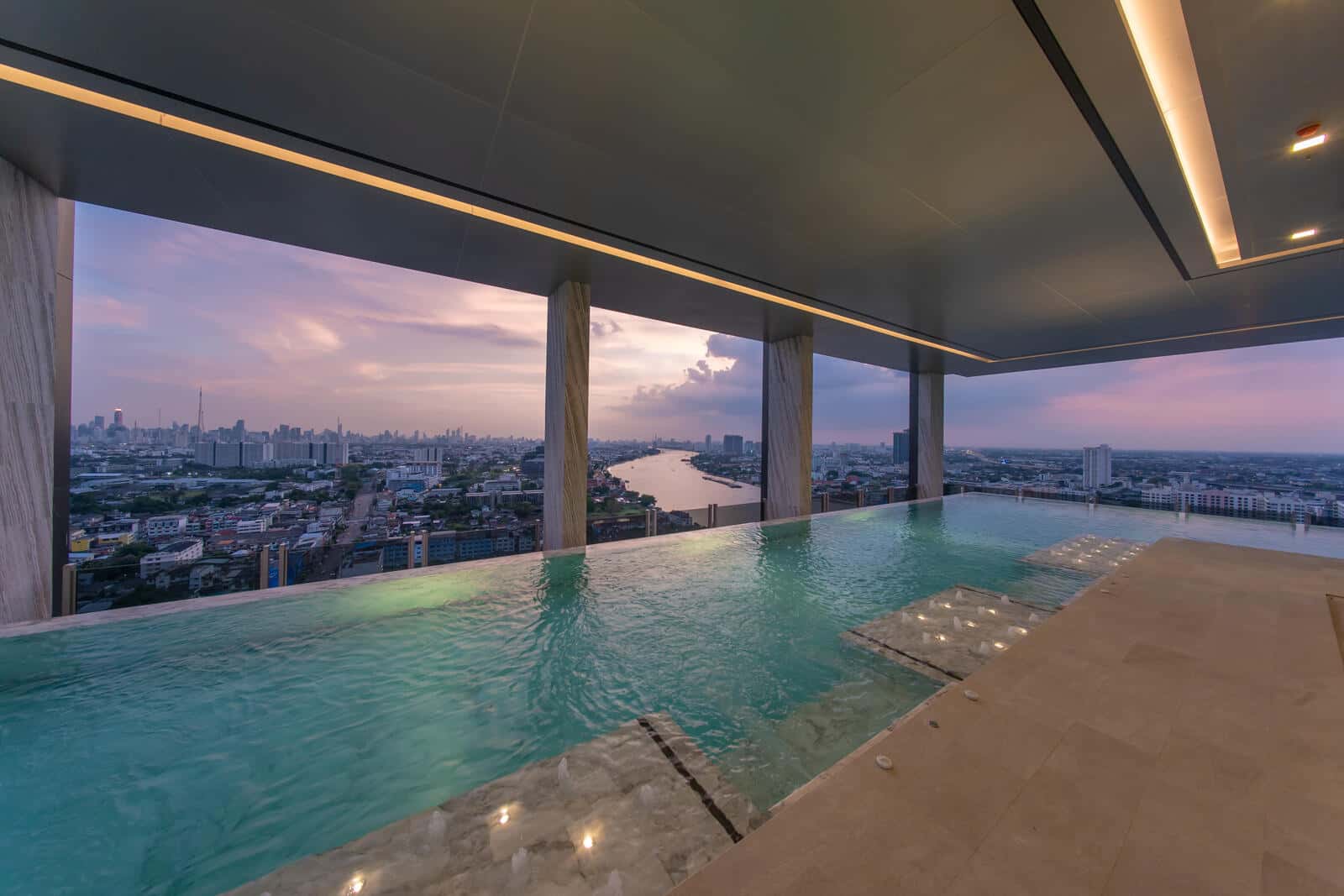Infinity-edge rooftop pool at Modiz Collection Bangpho at dusk, looking between stone columns over the Chao Phraya River and the Bangkok skyline