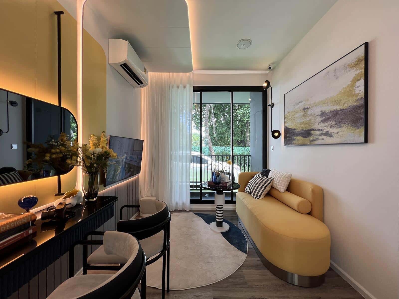 Show unit living area at Modiz Vault Kaset-Sripatum with a curved mustard sofa, a black console under a backlit mirror and a glazed door opening onto the balcony over mature trees