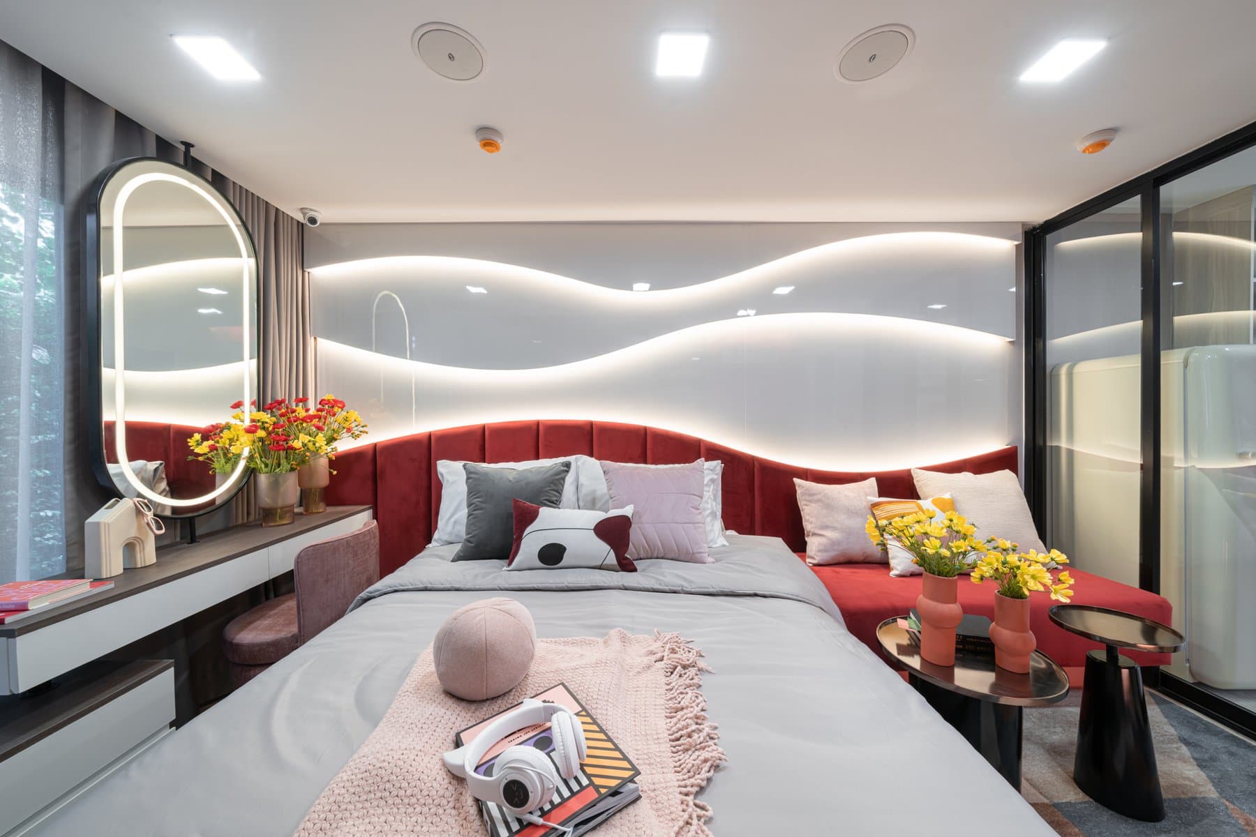 Bedroom of a furnished show unit at Atmoz Flow Minburi, with a deep red upholstered headboard, an oval backlit mirror and a sculpted wave motif on the wall