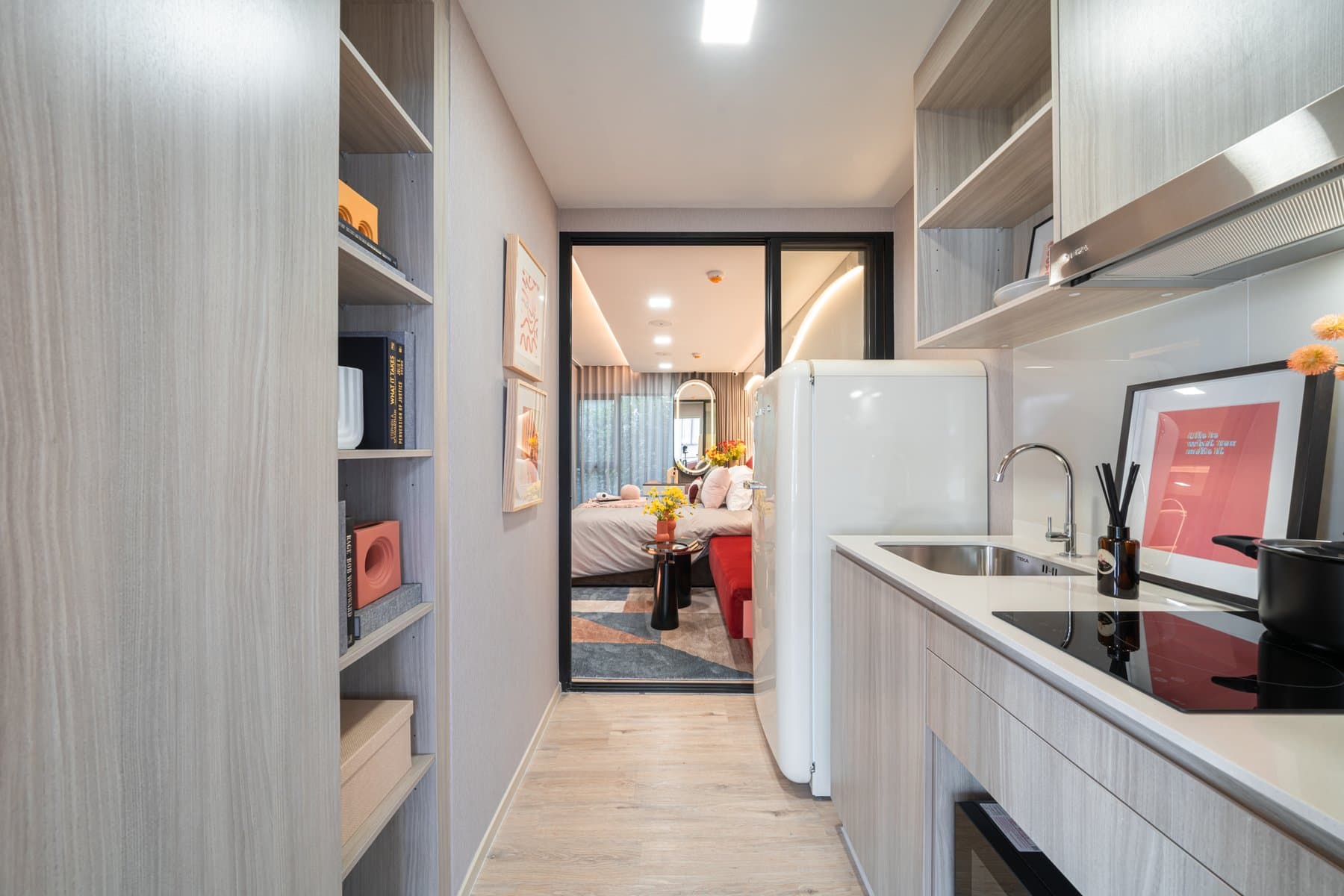 Galley kitchen in a furnished show unit at Atmoz Flow Minburi, with pale timber cabinetry, a built-in oven and a tall fridge, looking through to the bedroom beyond