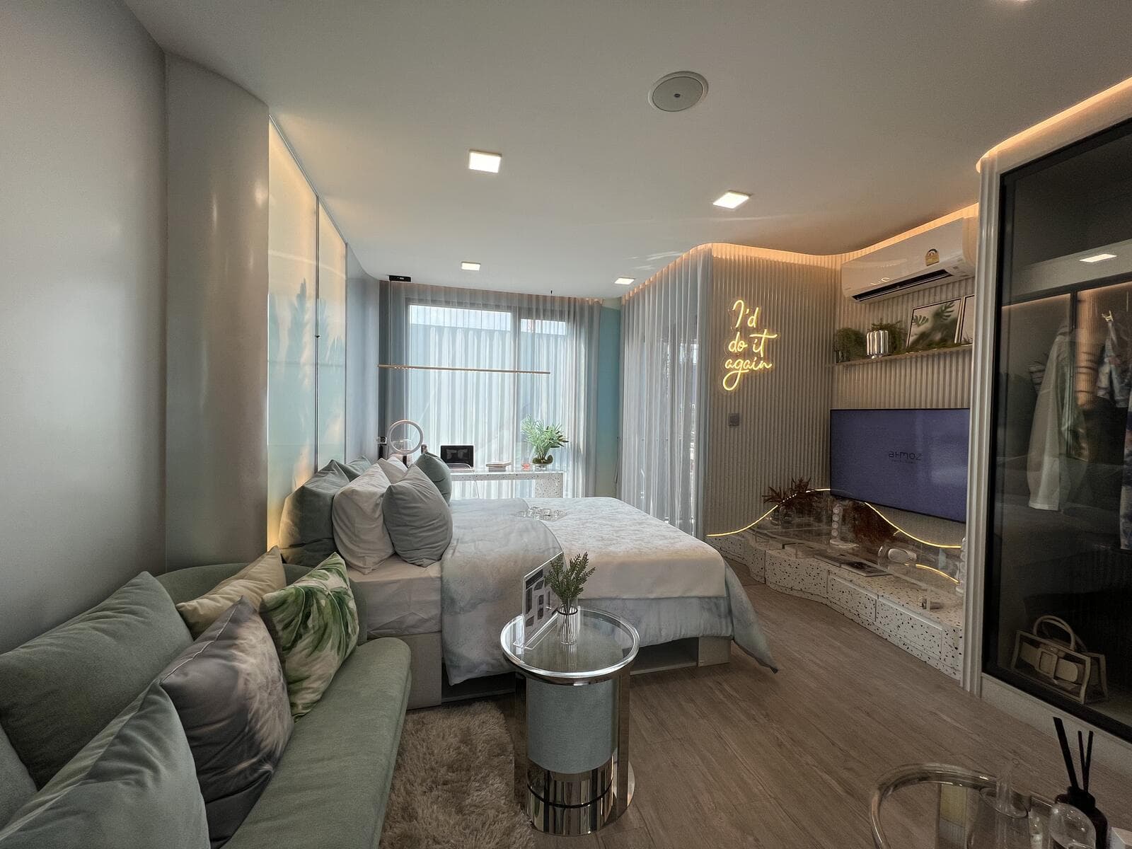 Show unit of a 24.10 sq.m. one-bedroom layout at Atmoz Oasis Onnut, with the bed, sofa, wall-mounted television and a full-height window behind sheer curtains