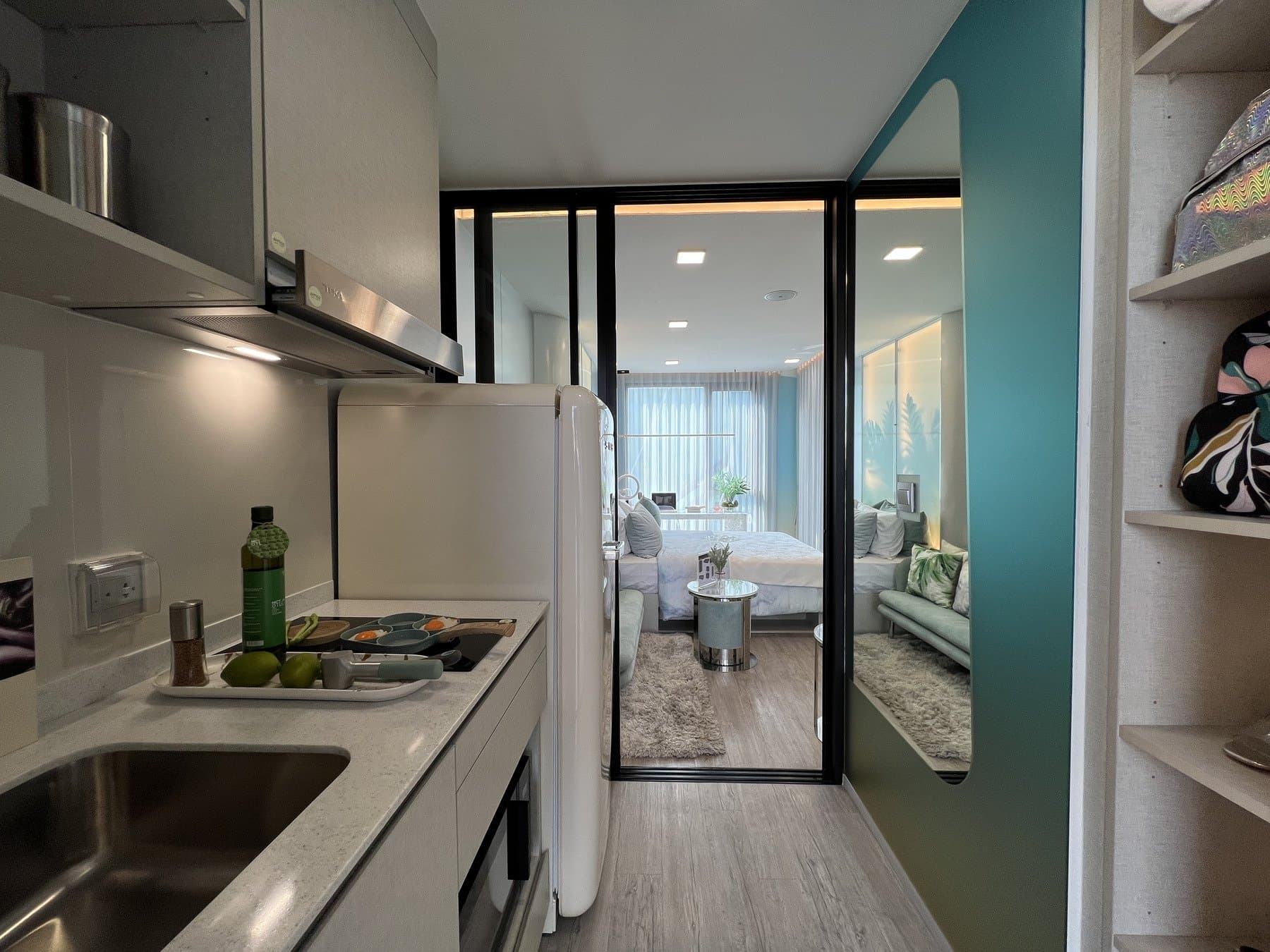 Kitchen of a furnished show unit at Atmoz Oasis Onnut, with a stainless sink, a black hob and open shelving, looking through the glazed partition to the bedroom