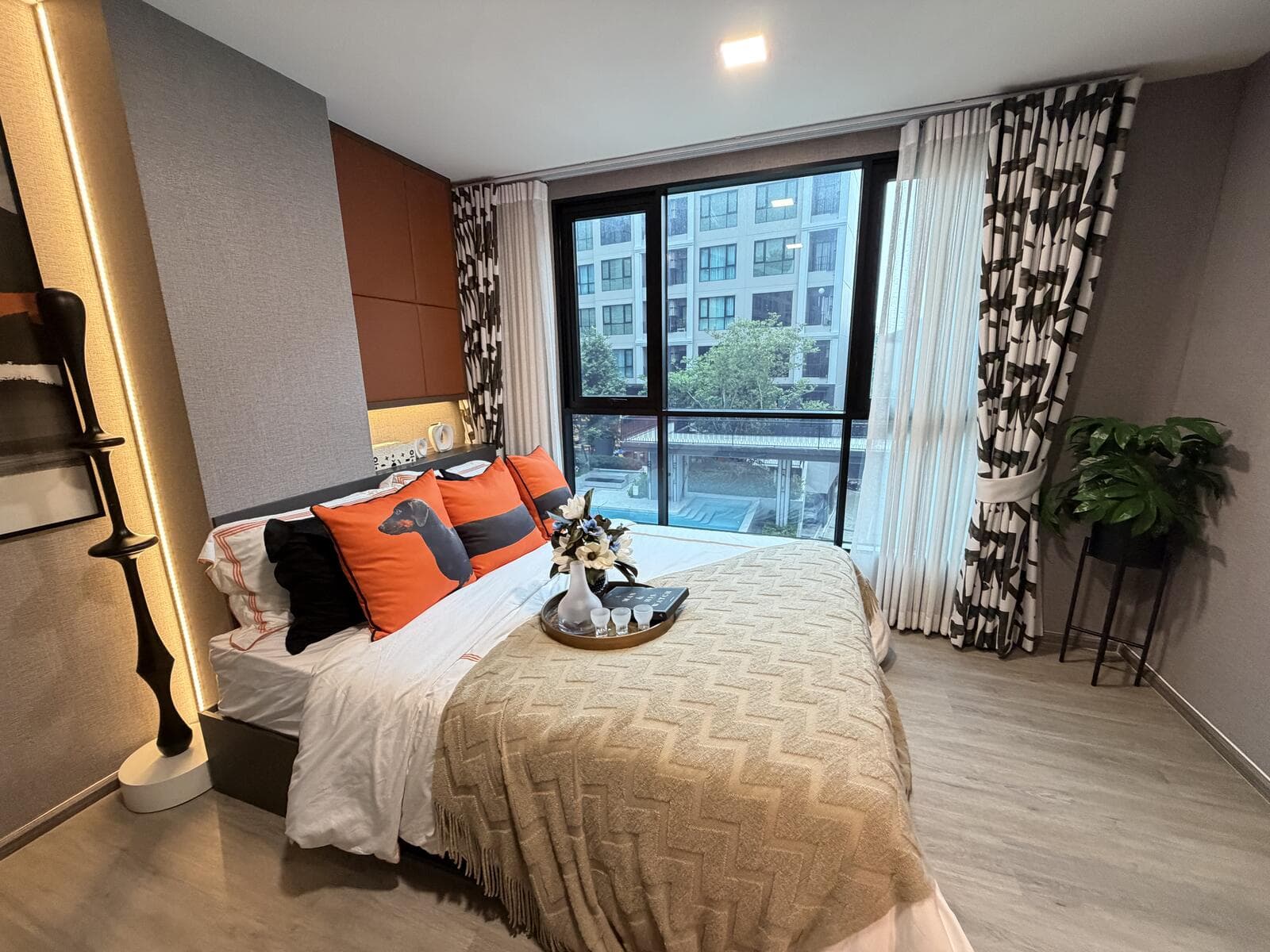 Show unit bedroom with a double bed against a padded headboard wall, patterned curtains and a floor-to-ceiling window overlooking the neighbouring residential wing.