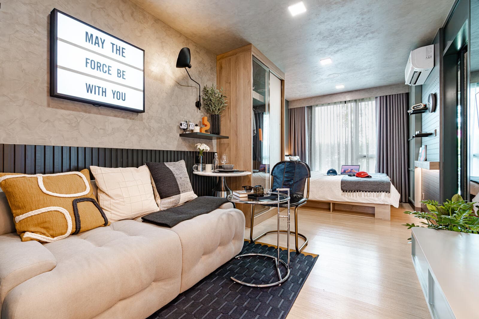 Show unit living area at Atmoz Season Ladkrabang, with a cream fabric sofa, glass side table and the sleeping area beside the curtained window