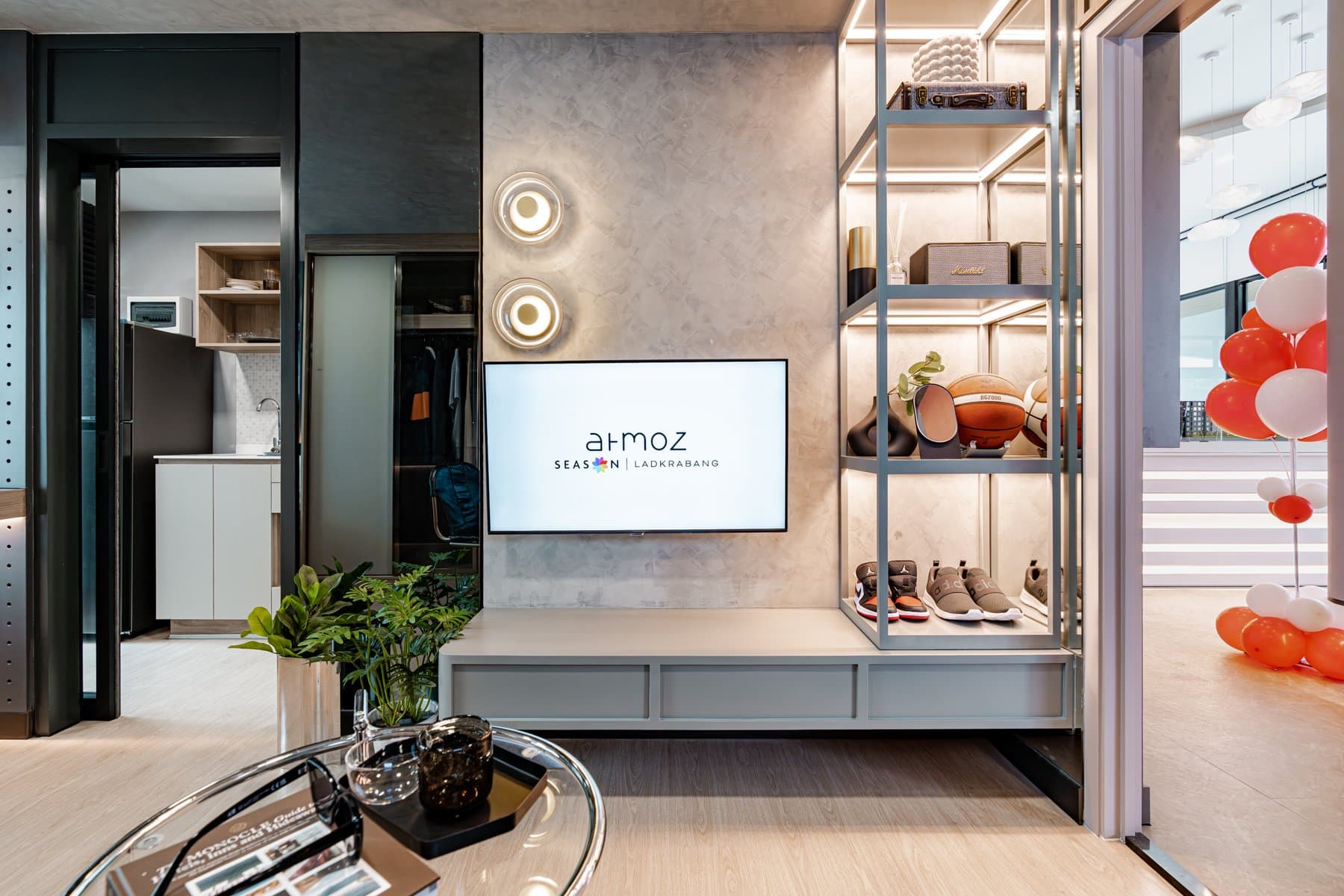 Living area of a furnished show unit at Atmoz Season Ladkrabang, with a wall-mounted television, open display shelving and a glass coffee table