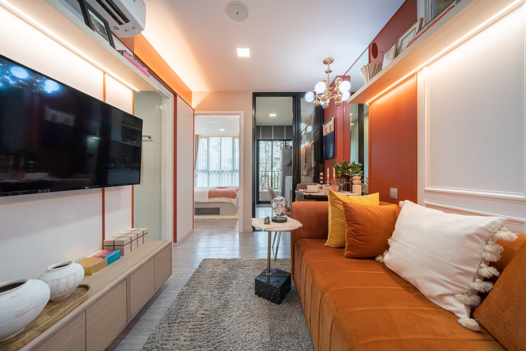 Living area of a furnished show unit at Atmoz Portrait Srisaman, with a burnt-orange sofa, a wall-mounted television and a brass pendant over the dining corner