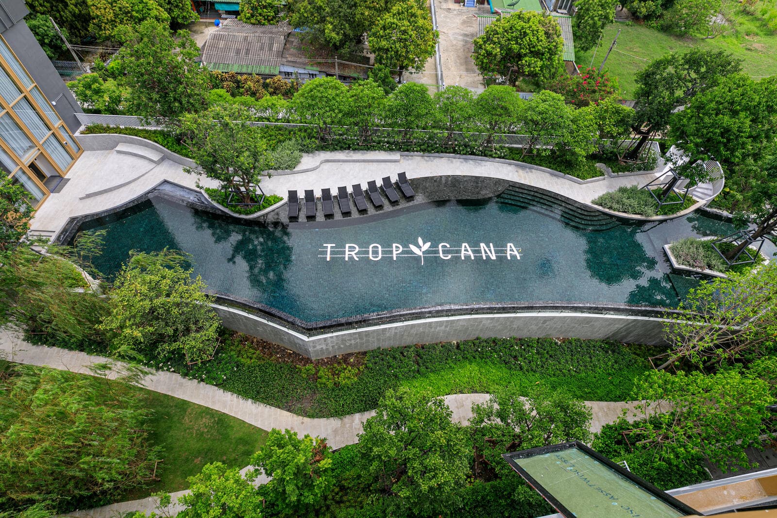 Aerial view of the curved outdoor swimming pool and sun deck at Atmoz Tropicana Bangna, surrounded by mature trees and garden paths