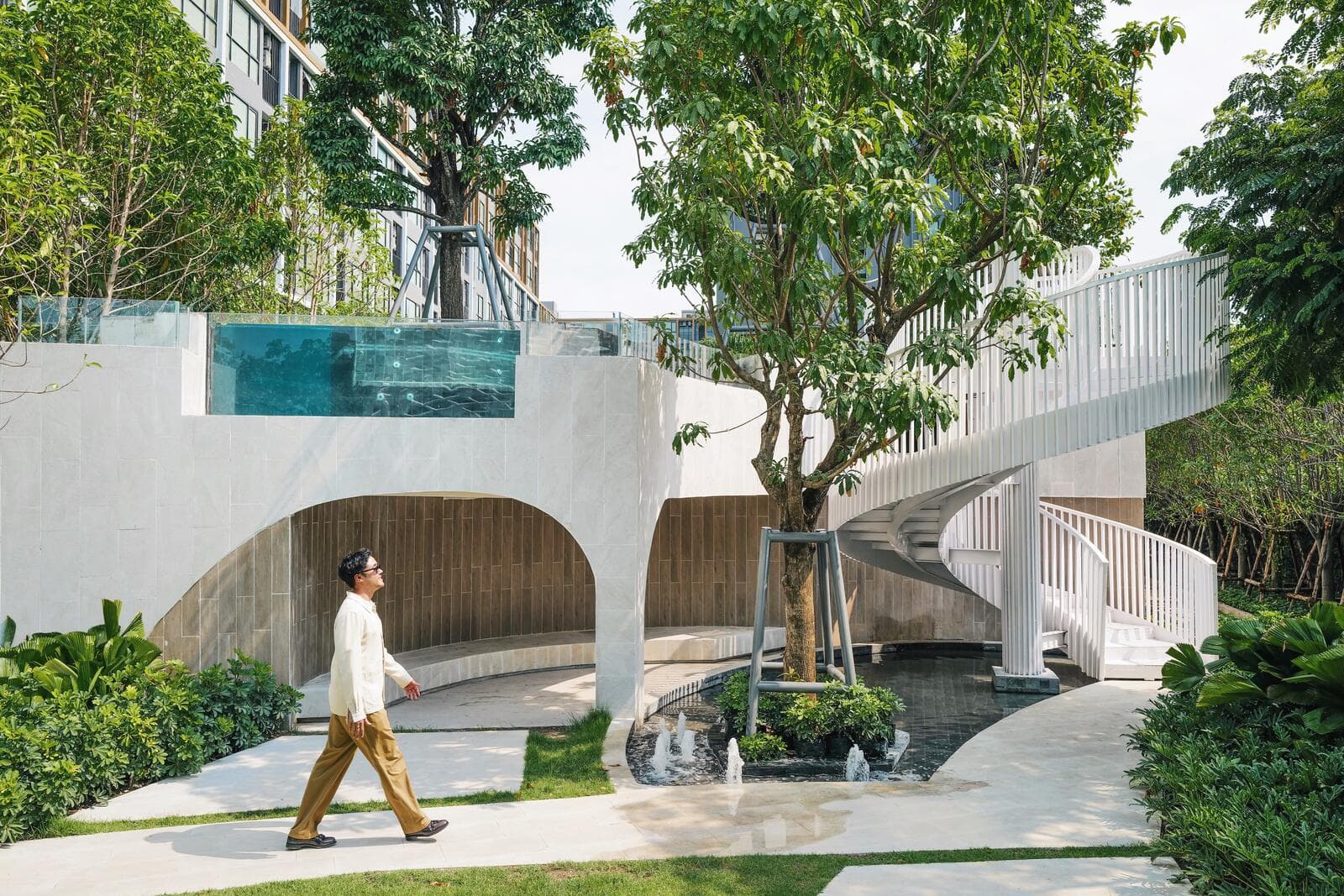 Garden courtyard at Atmoz Tropicana Bangna with an elevated glass-walled pool, arched shade wall, spiral stair and water feature