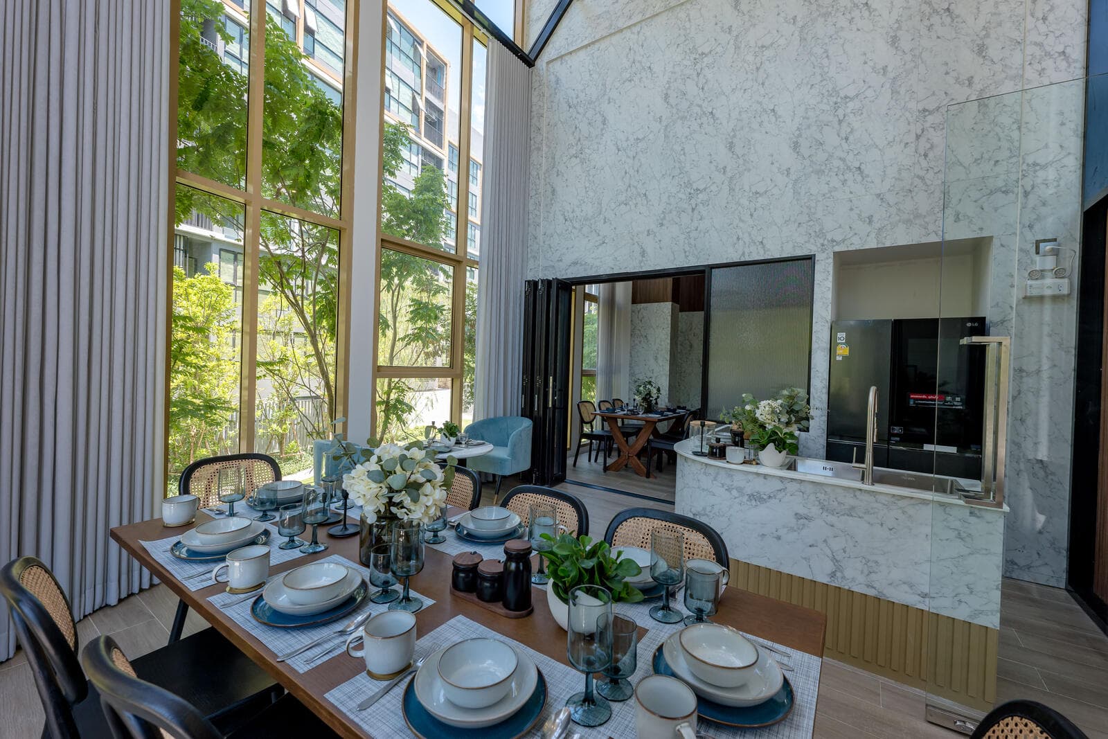 Double-height communal dining room and shared kitchen at Atmoz Tropicana Bangna with a long timber table, marble island and full-height garden windows