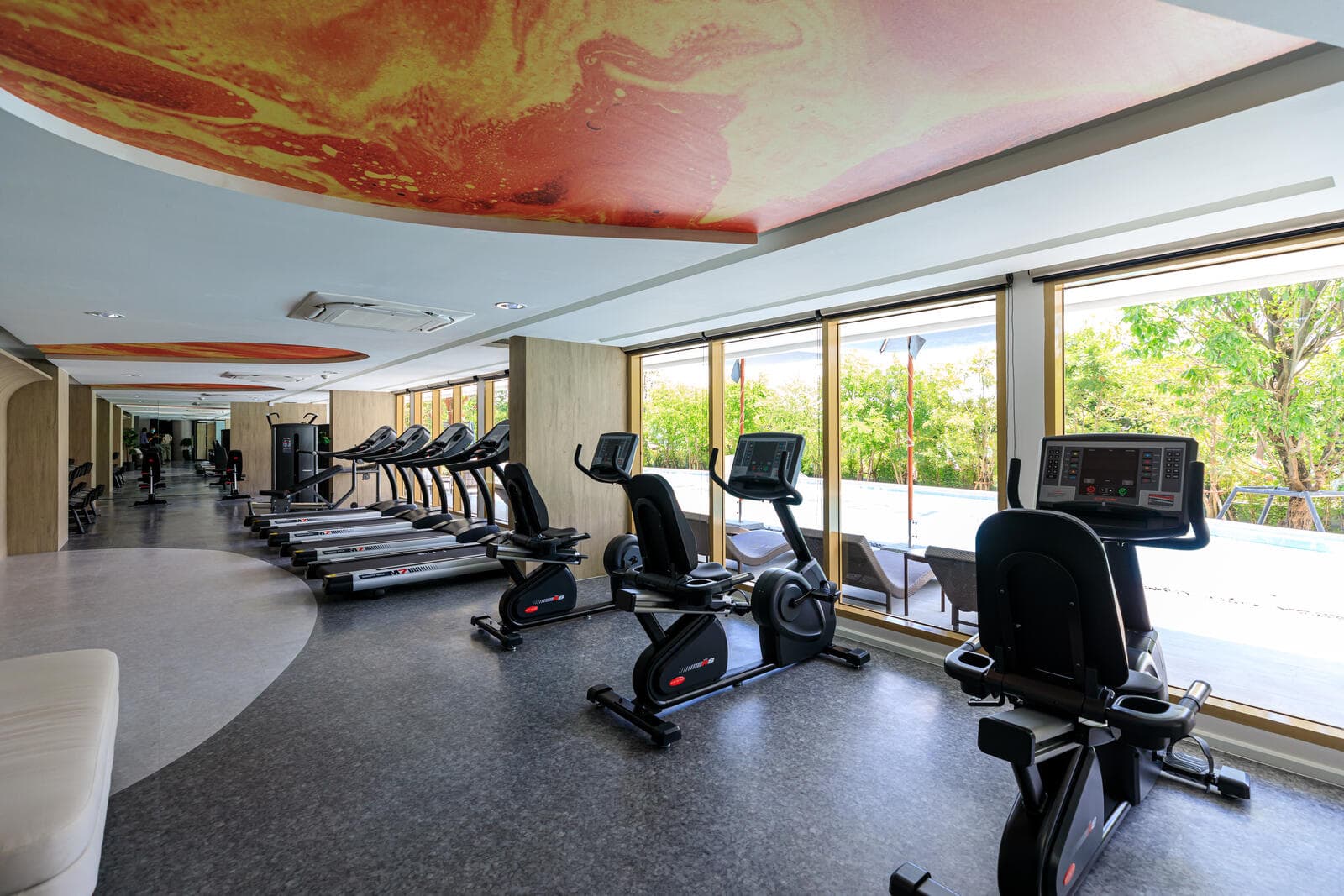 Fitness room at Atmoz Tropicana Bangna with treadmills and exercise bikes along a glazed wall overlooking the pool and garden