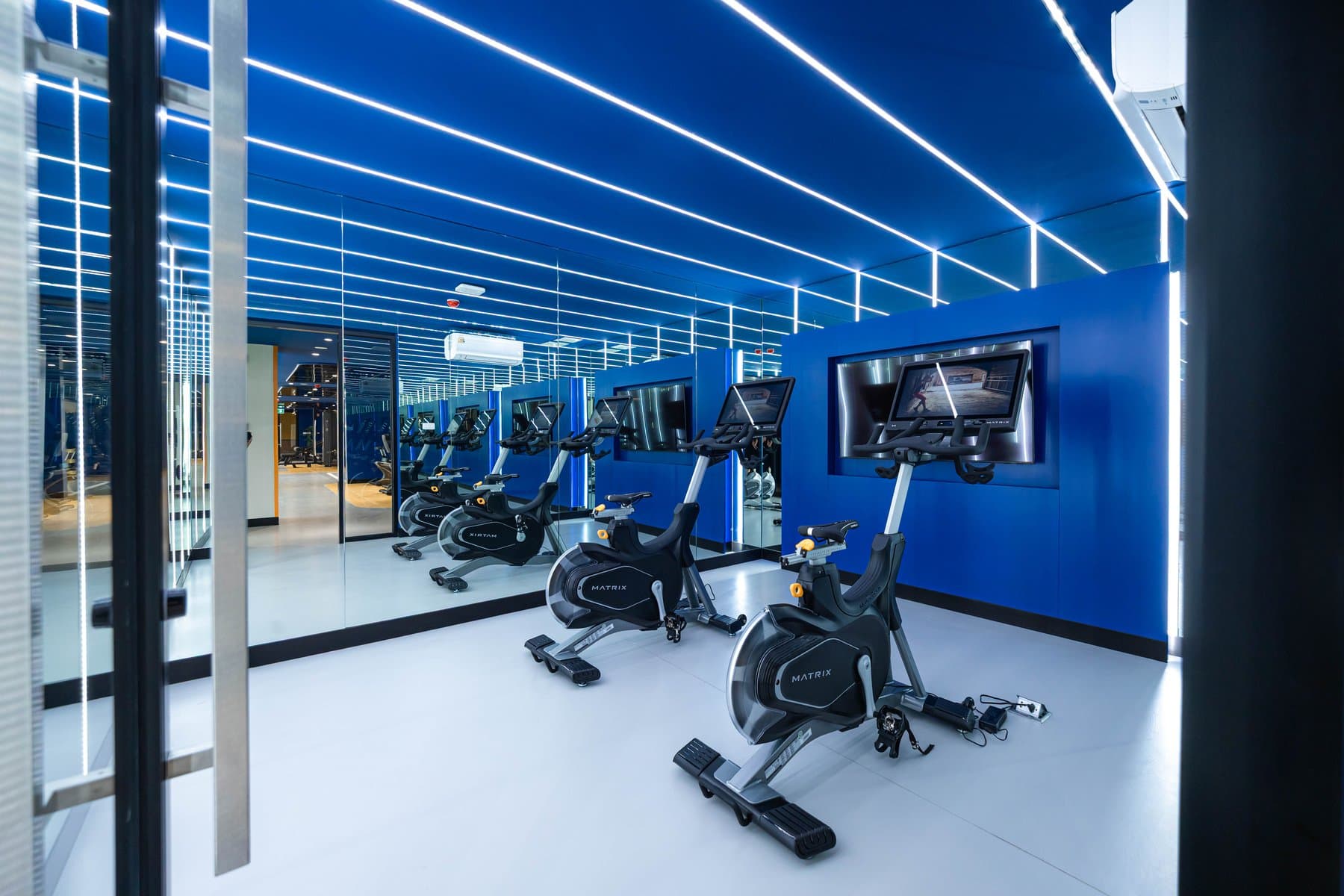 Fitness room at Kave COCO Bangsaen with exercise bikes, a mirrored wall and blue linear ceiling lighting