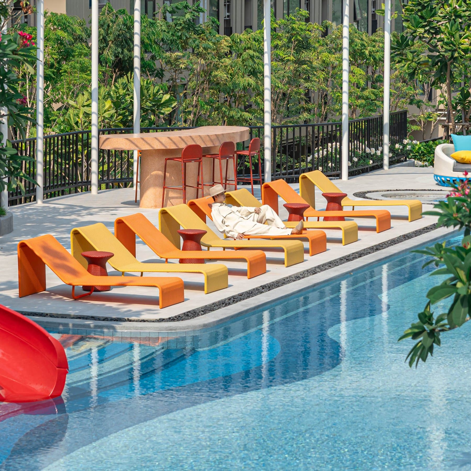 Swimming pool at Kave COCO Bangsaen with a row of orange sun loungers along the deck and mature trees behind