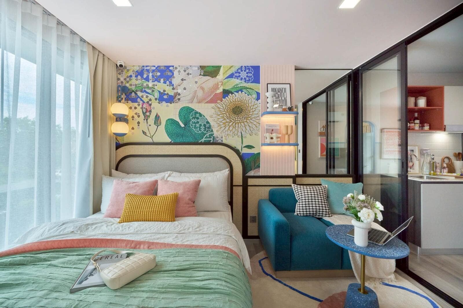 One-bedroom show unit: bed against a patterned feature wall, teal sofa and side table, with the fitted kitchen visible through the glass partition