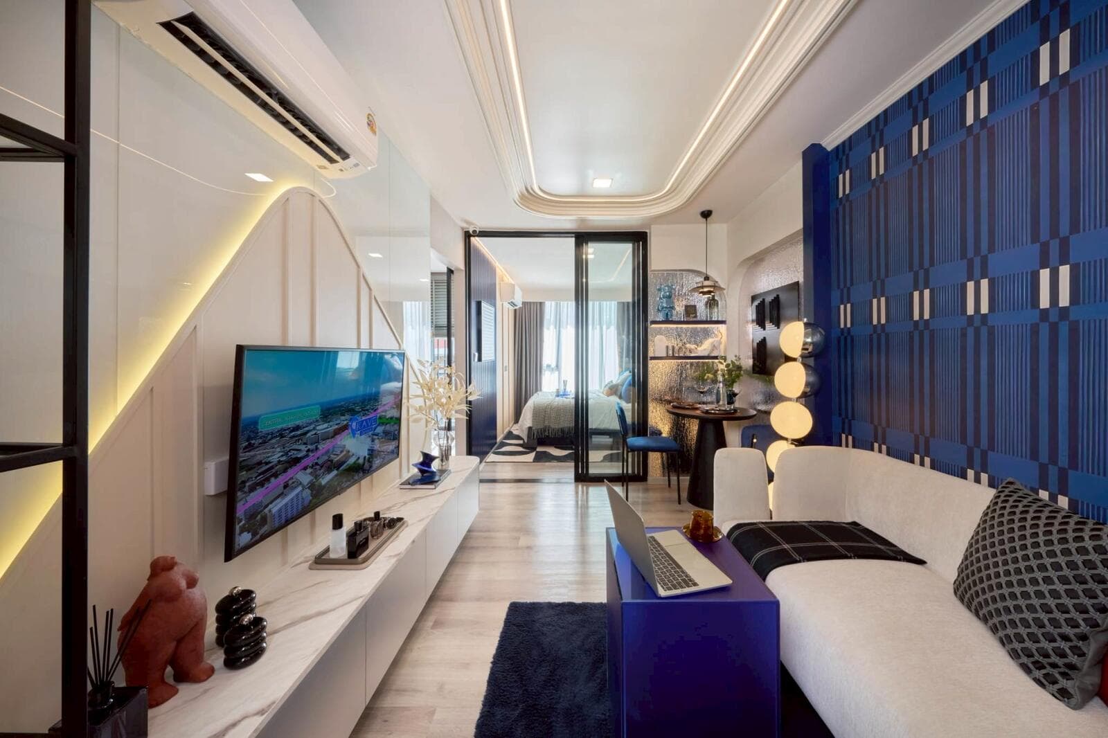 One Bedroom Exclusive show unit living area with wall-mounted television on a marble-topped console, sofa, dining nook and the bedroom beyond the sliding glass door