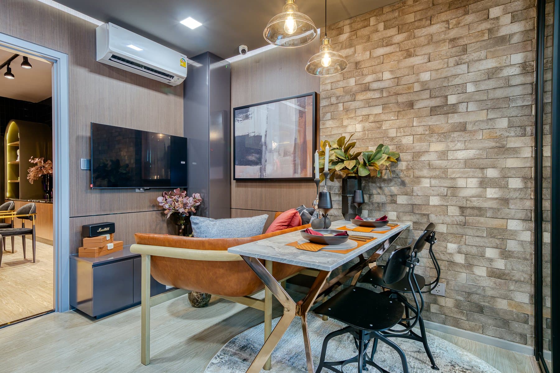 Dining table in a furnished show unit at Maroon Ratchada 32, set beneath pendant lights against an exposed brick-effect wall with the television alongside