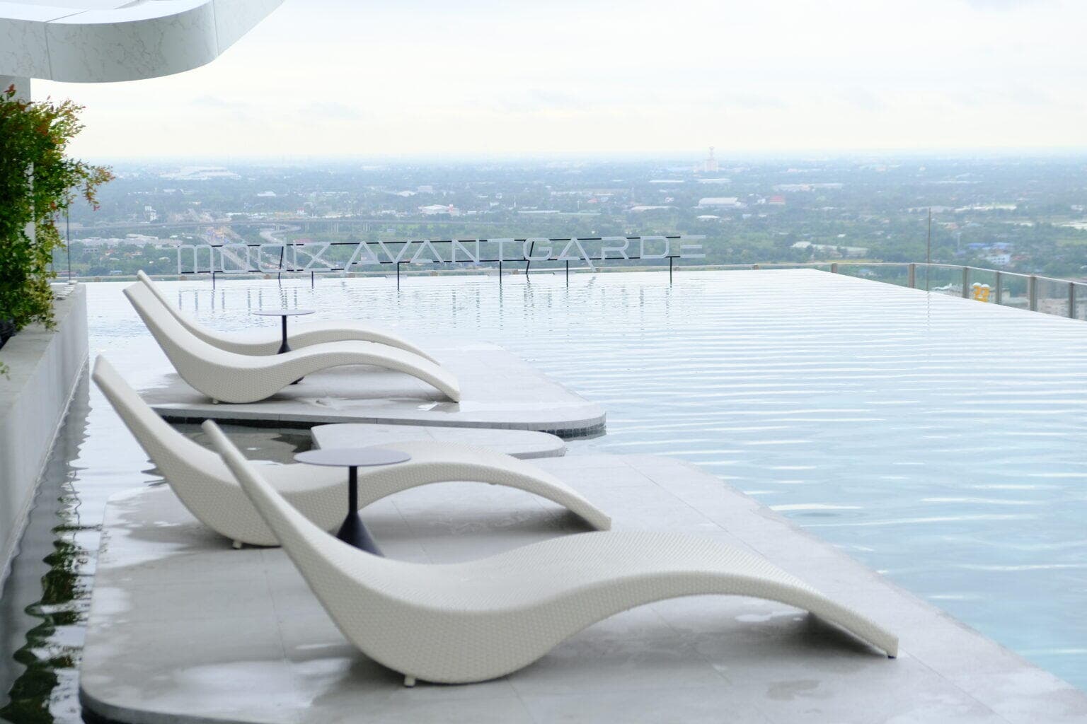 Rooftop infinity-edge swimming pool at Modiz Avantgarde with white contour sun loungers and side tables, the project lettering standing in the water and the Khlong Luang plain stretching to the horizon