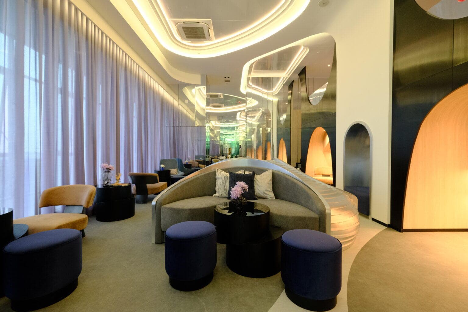 Residents' lounge at Modiz Avantgarde with curved cove lighting, sheer full-height curtains, a rounded banquette sofa, velvet drum stools and mirrored arch alcoves