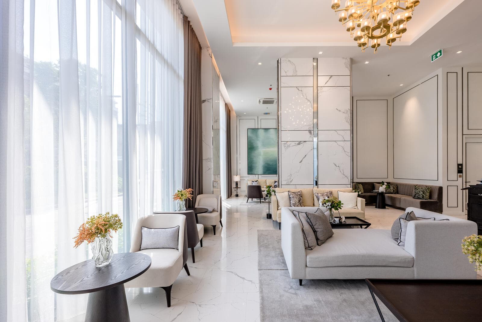 Ground-floor residents' lounge at Modiz Collection Bangpho with double-height sheer-curtained glazing, marble wall panels, a brass ring chandelier and pale upholstered seating on white marble flooring
