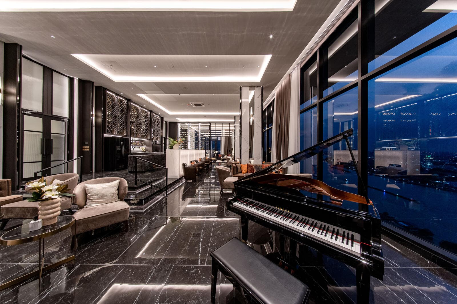 Sky lounge and wine bar at Modiz Collection Bangpho at night, with a grand piano, polished black marble flooring, backlit metal screens and full-height glazing looking over the river