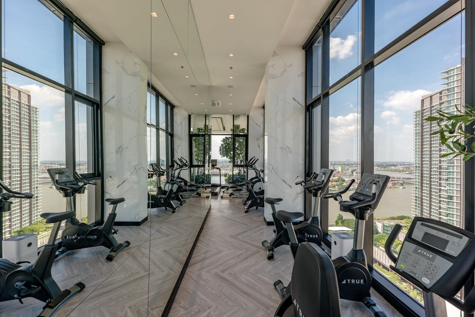 Sky fitness room at Modiz Collection Bangpho with upright exercise bikes, herringbone flooring, marble-clad columns, a mirrored wall and full-height windows over the Chao Phraya River