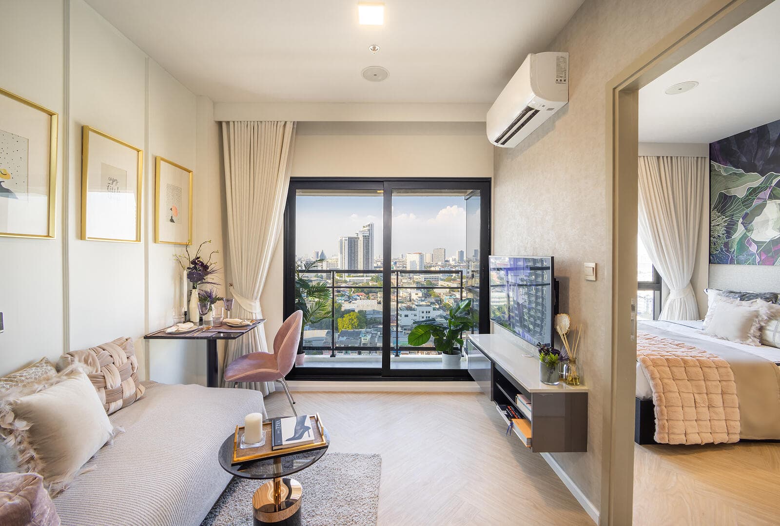 One-bedroom show unit at Modiz Collection Bangpho, showing the living area with a day bed, wall-mounted television console, compact dining table beside the balcony door and the bedroom through the opening