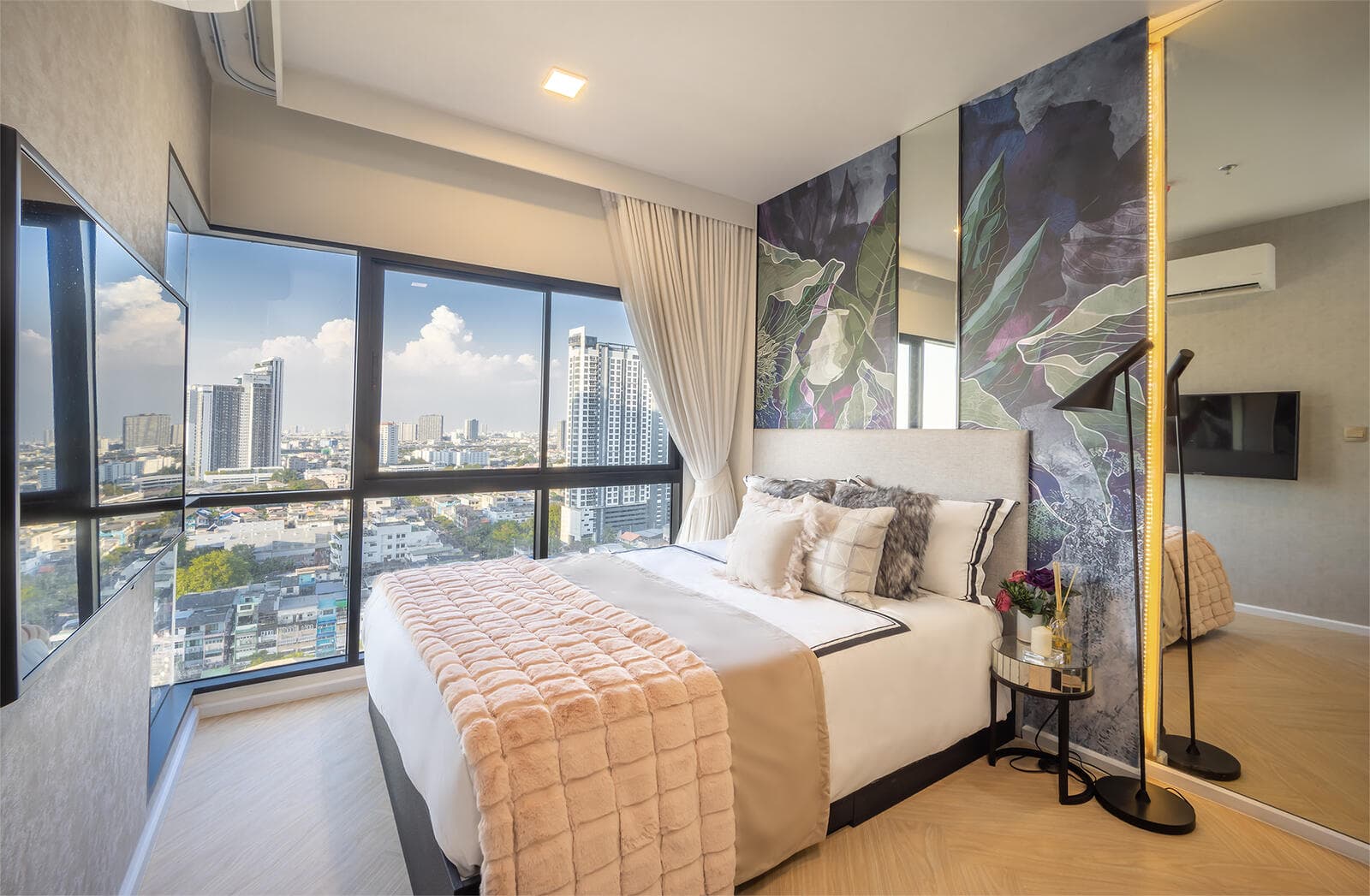 Bedroom in the one-bedroom show unit at Modiz Collection Bangpho, with an upholstered headboard against a botanical mural panel, wall-mounted television and corner windows onto the city