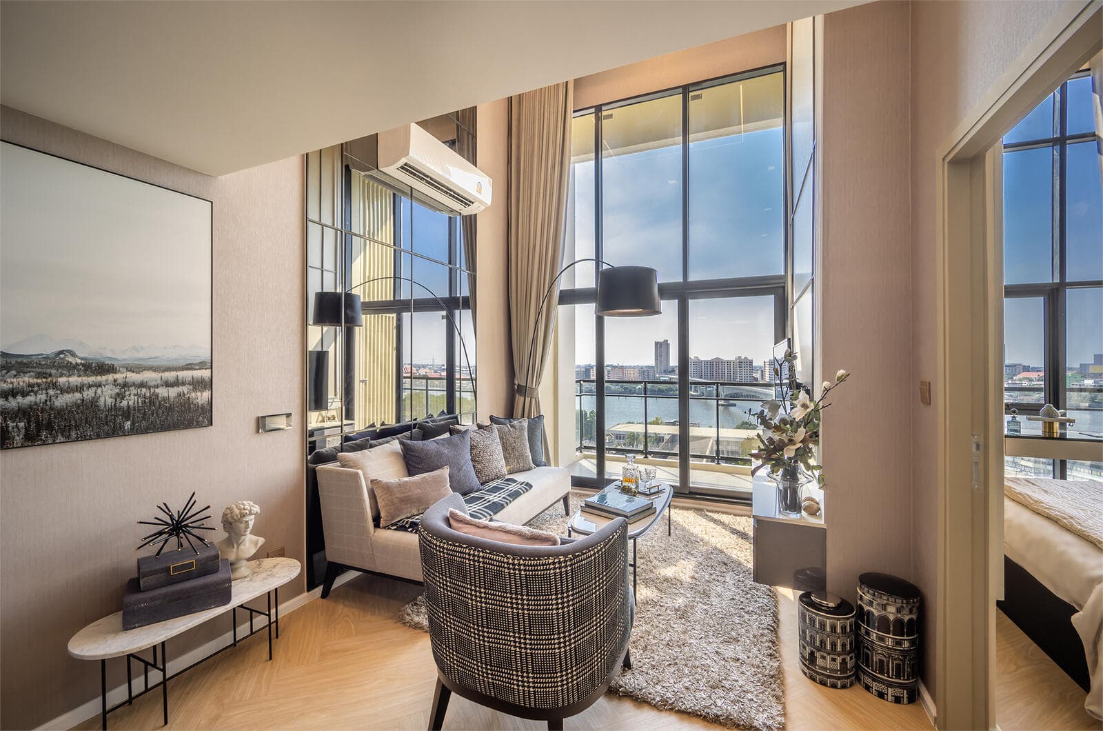 Living area of the two-bedroom vertical suite show unit at Modiz Collection Bangpho, with full-height glazing framing the Chao Phraya River, an arc floor lamp, mirrored feature wall and light timber-tone flooring