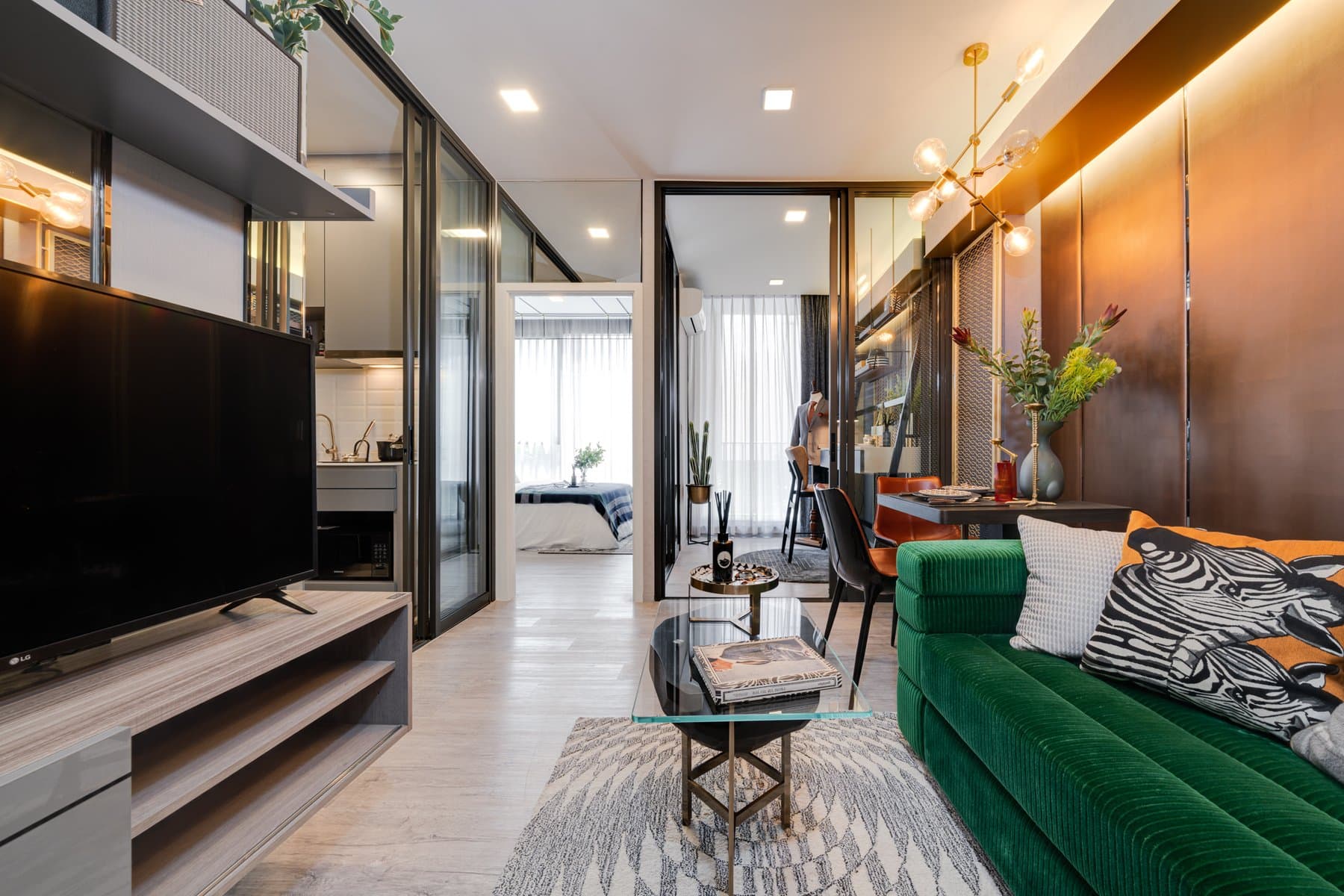 Living area of a furnished show unit at Modiz Rhyme Ramkhamhaeng, with an emerald velvet sofa, a television on the timber wall and a zebra-print stool