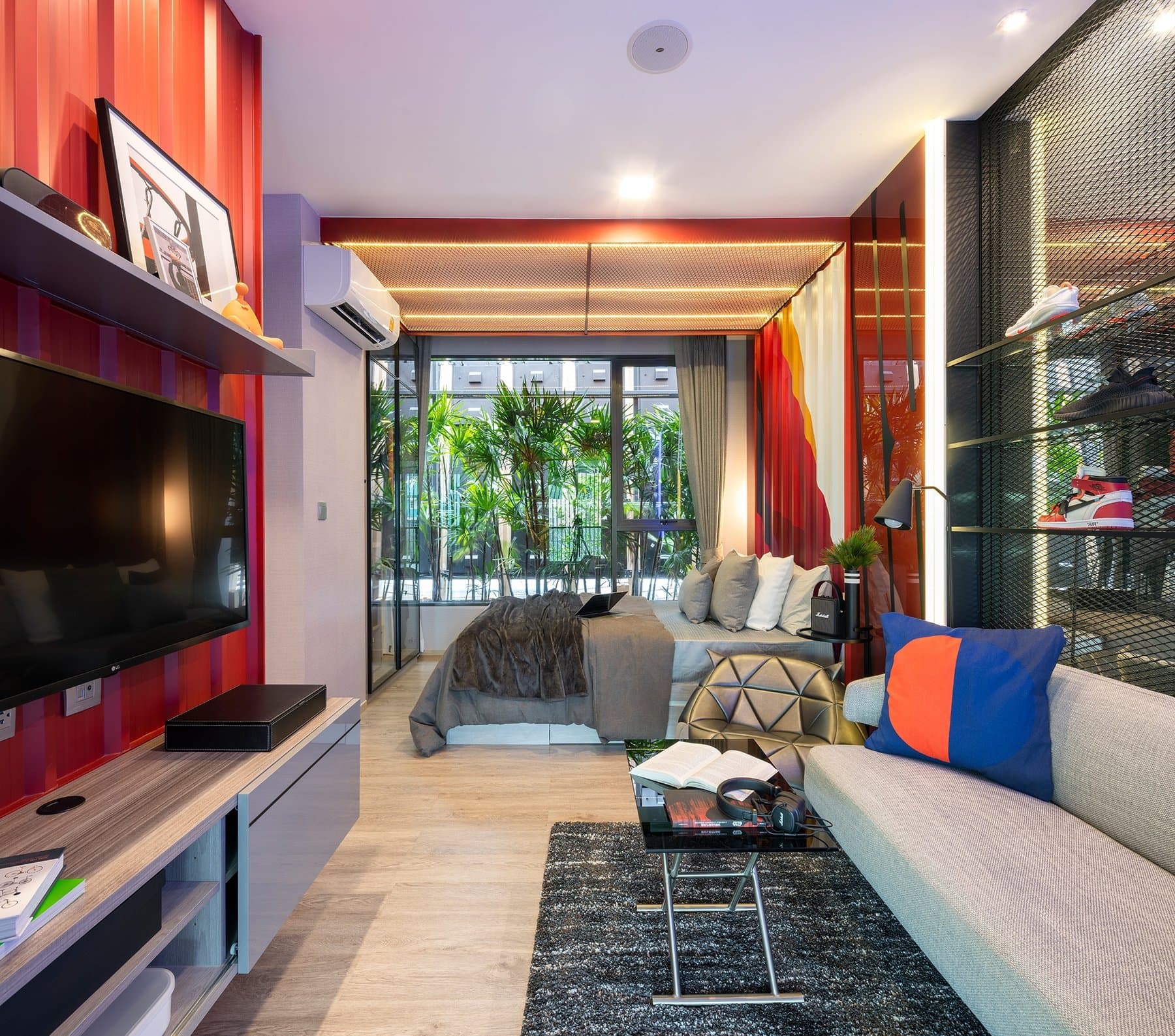 Living area of a furnished show unit at Modiz Rhyme Ramkhamhaeng, with a red feature wall, a wall-mounted television and a glazed screen to the balcony
