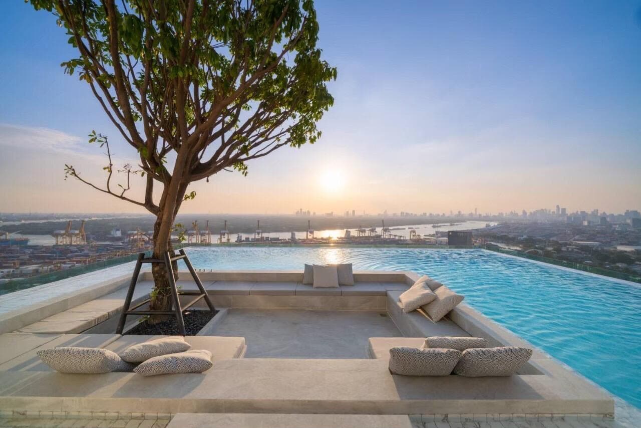 Rooftop infinity pool at Modiz Sukhumvit 50 at sunset, with a sunken sundeck, cushioned bench seating and a single tree framing the view across the river toward the Bangkok skyline