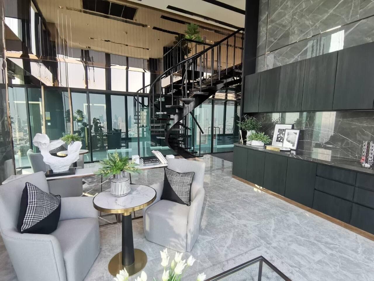 Double-height residents' lounge at Modiz Sukhumvit 50 with a black spiral staircase, marble flooring, grey armchairs and a glazed wall opening onto the fitness room and city view