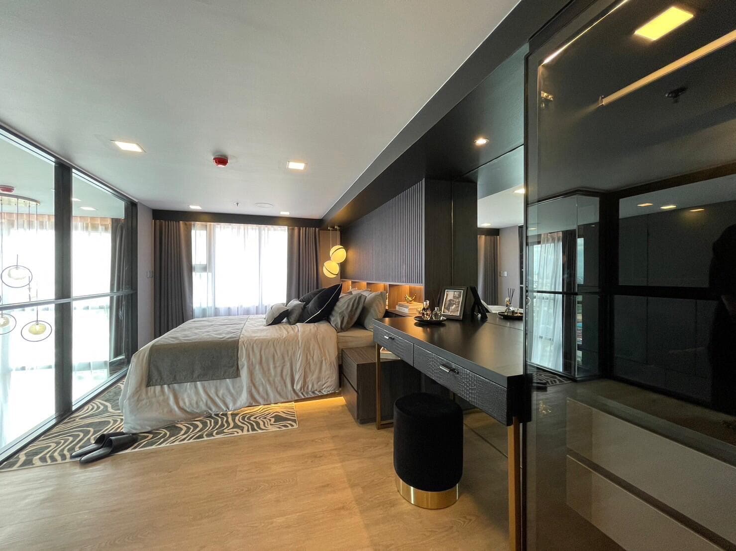 Show unit bedroom at Modiz Sukhumvit 50 on the mezzanine level, with a fluted dark headboard wall, brass pendant lights, a long dressing desk and a curtained window wall