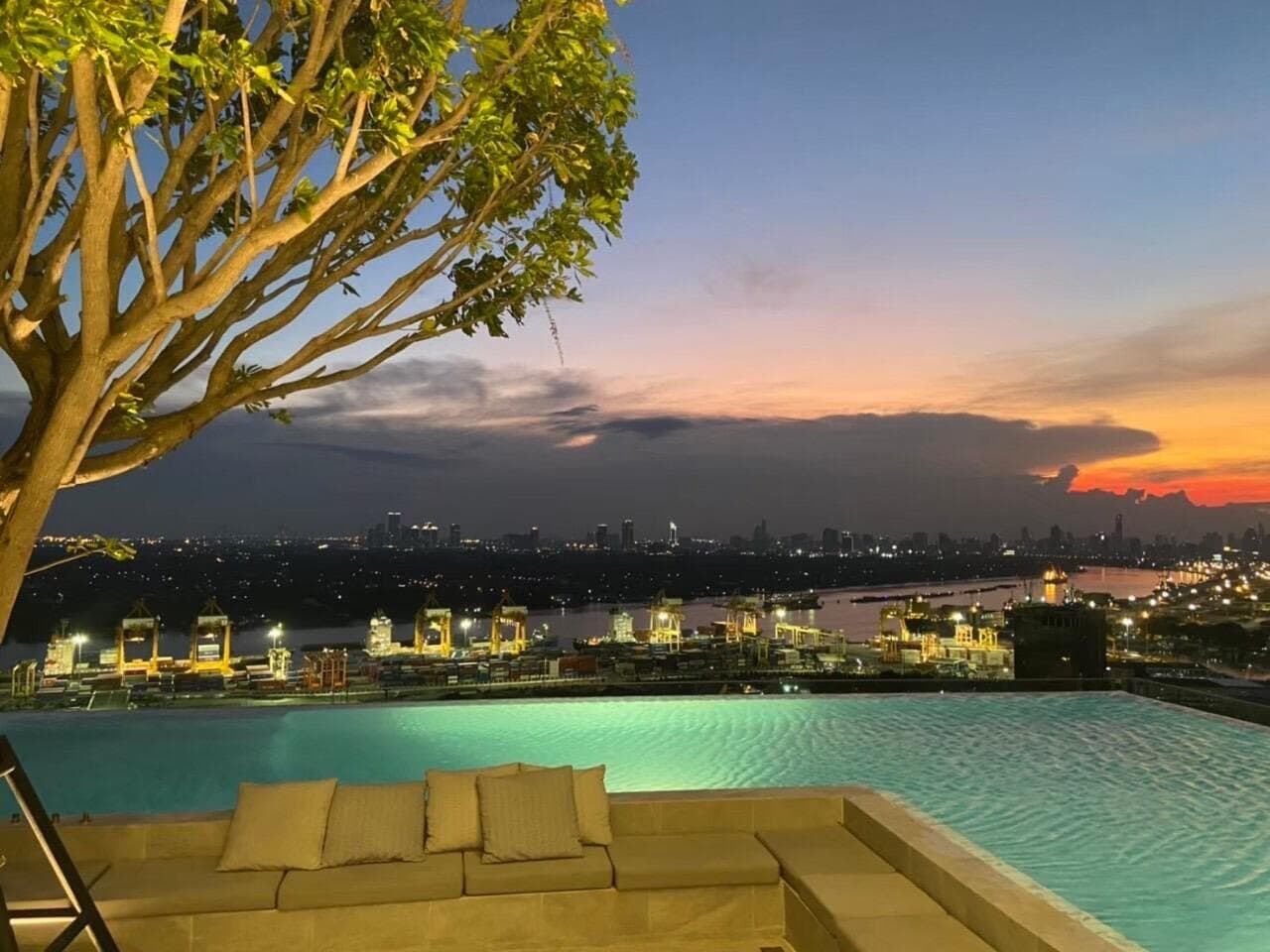 Evening view from the rooftop pool deck at Modiz Sukhumvit 50, looking past the lit pool edge over the port cranes and the river toward the Bangkok skyline at dusk