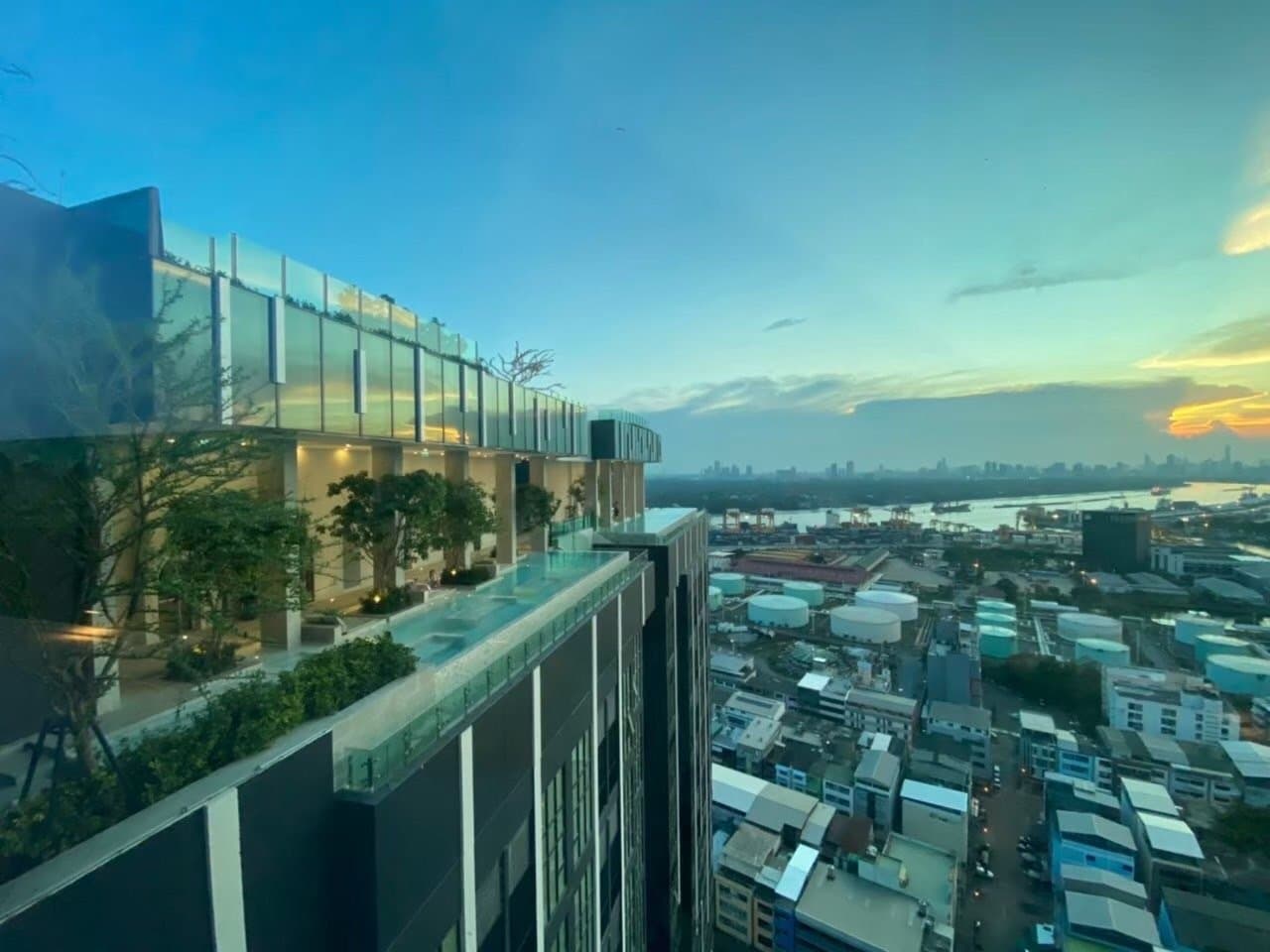 View from a balcony at Modiz Sukhumvit 50 at dusk, looking across neighbouring rooftops with the building's glazed corner in the foreground