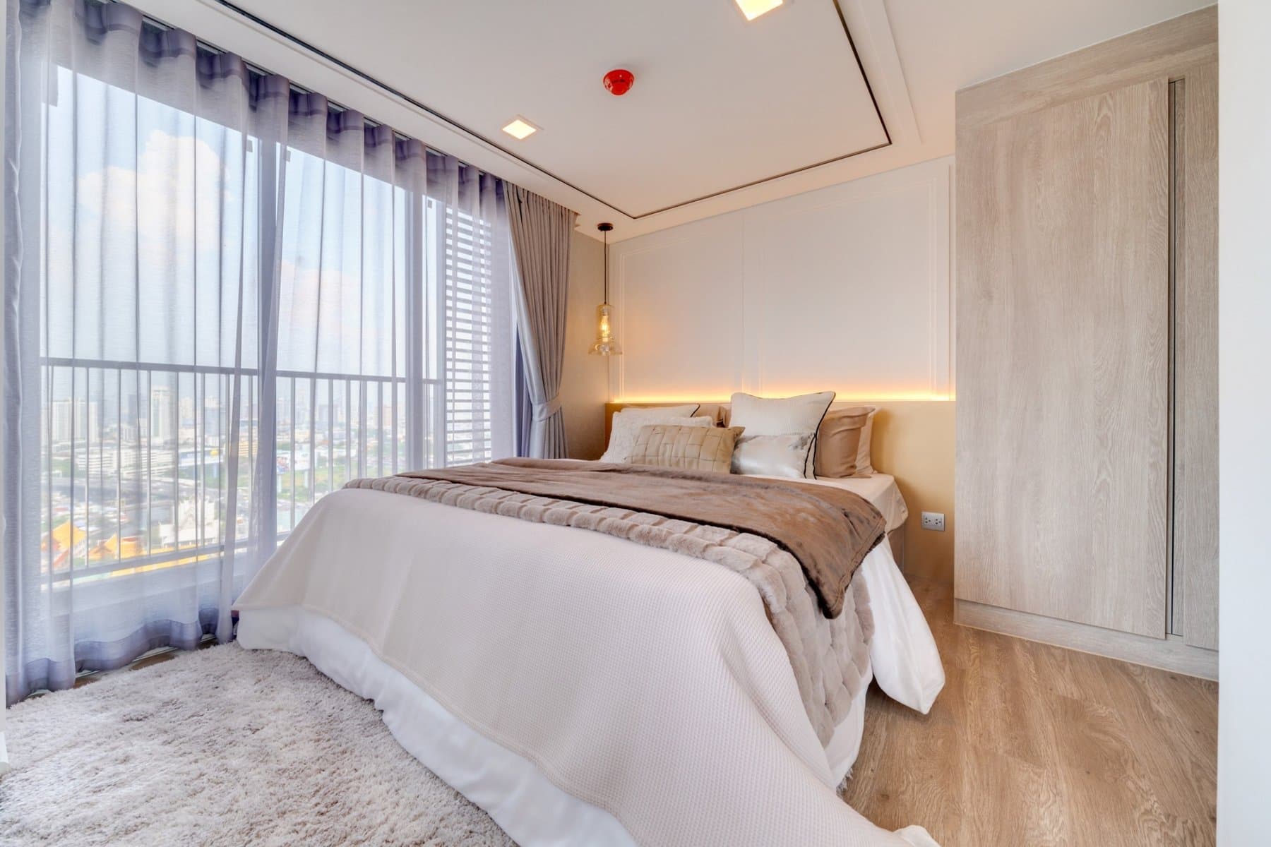 Bedroom of a furnished show unit at Modiz Sukhumvit 50, with layered cream bedding, a timber wardrobe and city daylight through the curtains