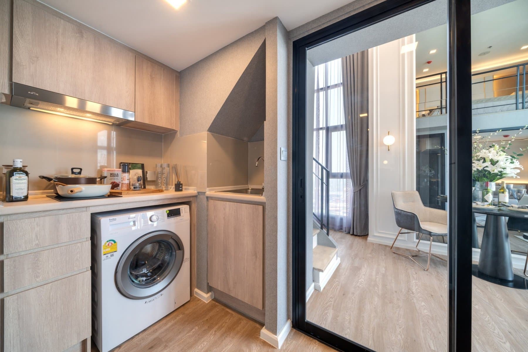 Kitchen of a furnished show unit at Modiz Sukhumvit 50, with a washing machine below the counter, overhead cupboards and a glazed partition