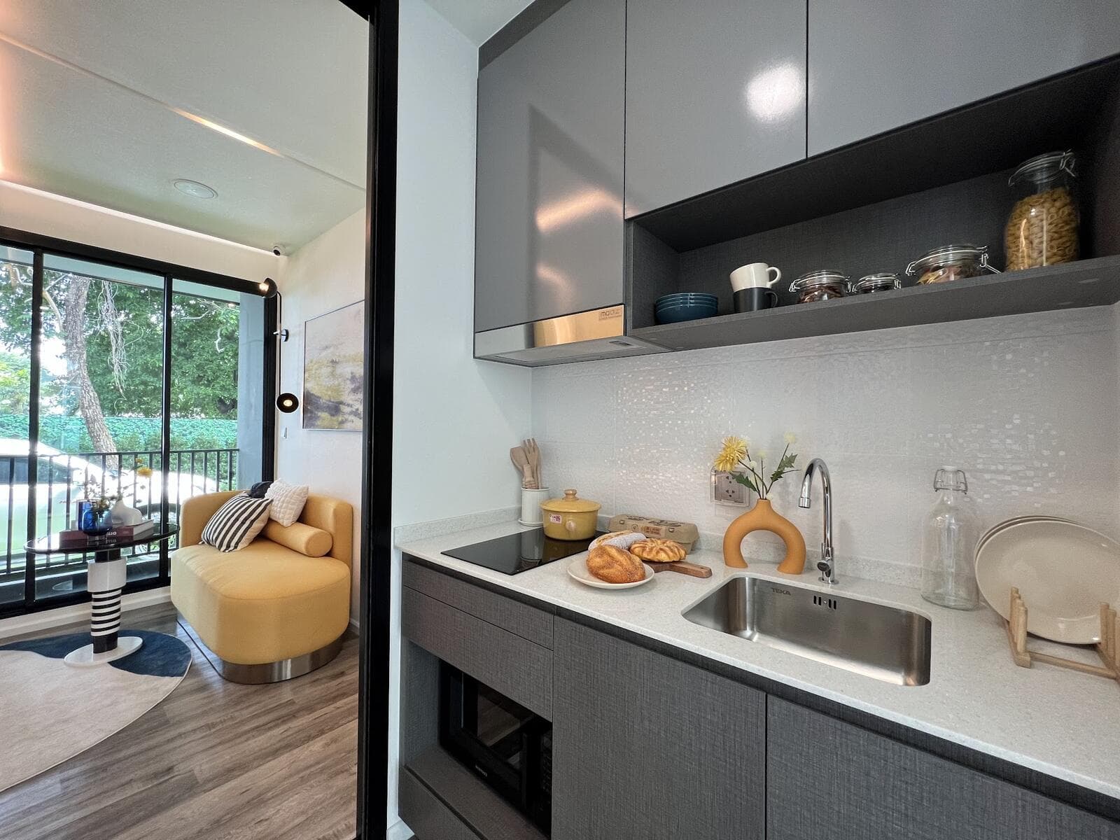 Show unit kitchen at Modiz Vault Kaset-Sripatum, 1 Bedroom Extra 28.10 sq.m: a fitted graphite galley with stone worktop, induction hob, undermount sink, extractor hood and built-in oven, looking through to the living area and balcony