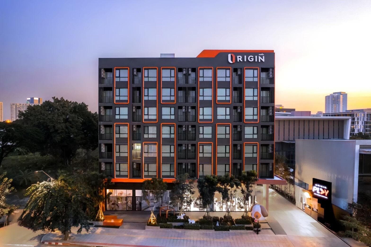 Photograph of the completed Origin Play Sri Udom Station building at dusk, showing the dark grey facade with orange window frames and the lit ground-floor common area.