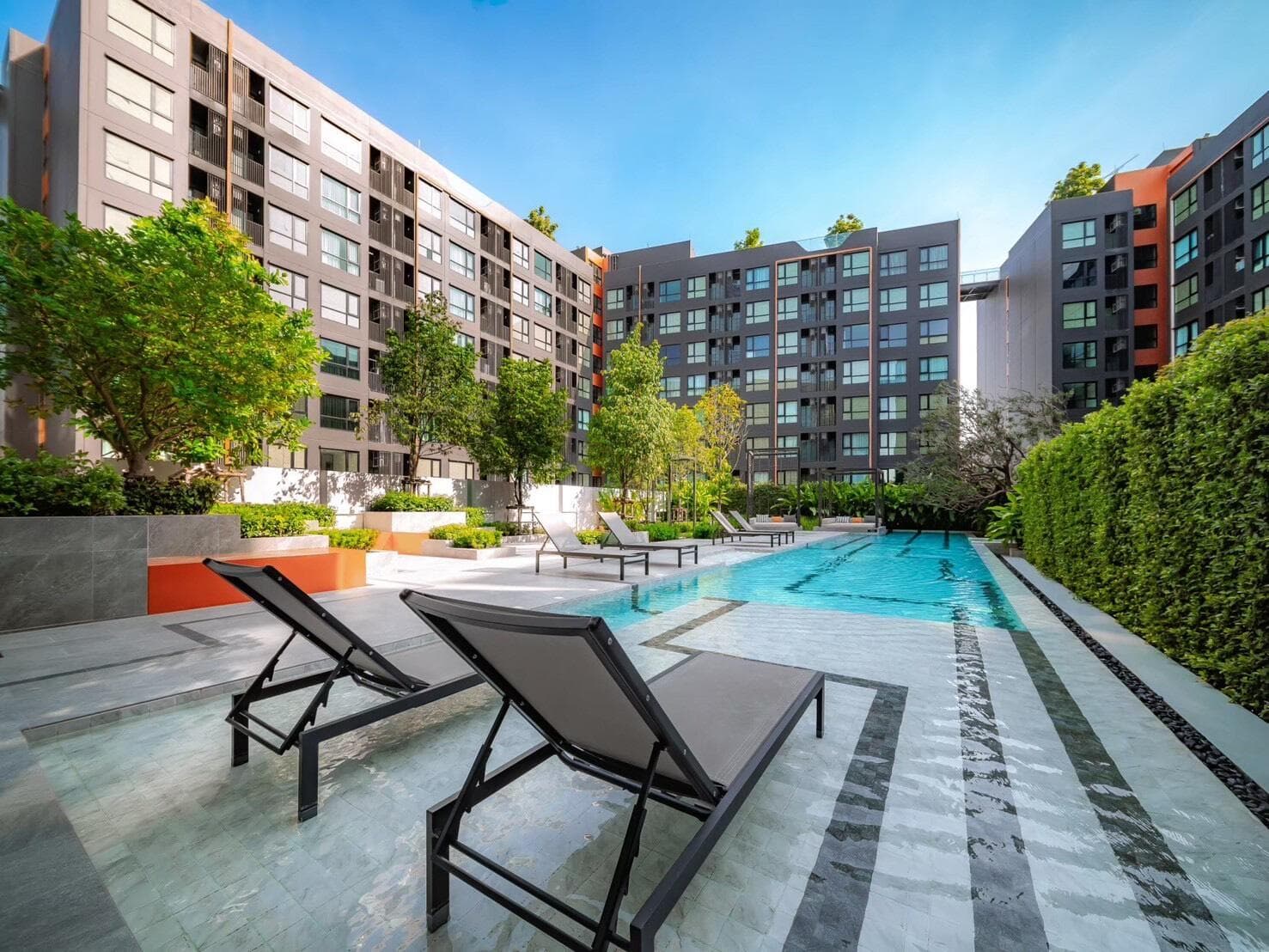 Photograph of the outdoor lap pool at Origin Play Sri Udom Station with sun loungers on the stone deck and the residential buildings around the landscaped courtyard.
