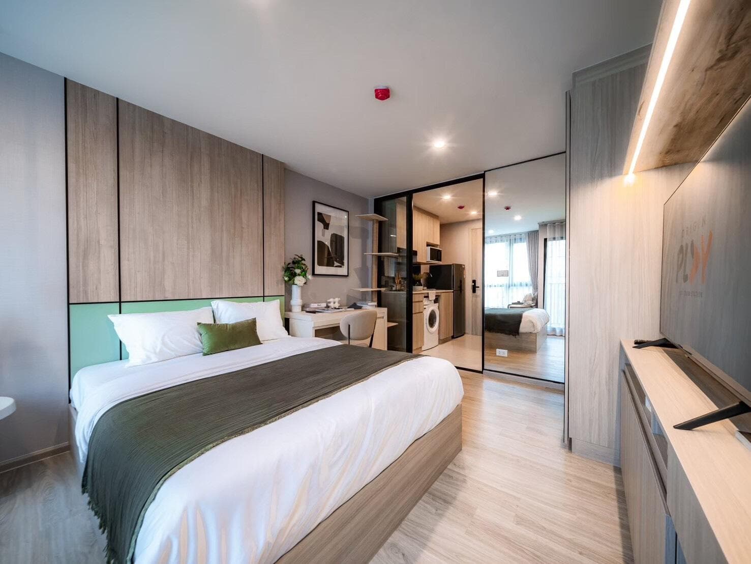Show unit photograph of a 24 sqm studio looking from the bed toward the glazed partition, kitchen counter and washing machine, with a wall-mounted television at right.