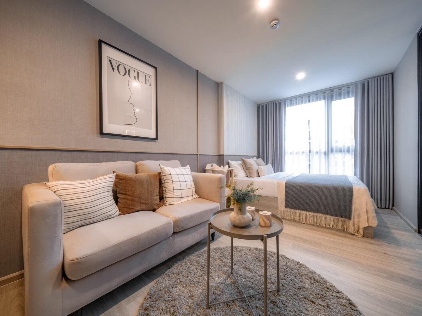 Show unit photograph of a 28 sqm Smart Closet unit with a fabric sofa and round side table in the living zone and the bed beside the full-height window.