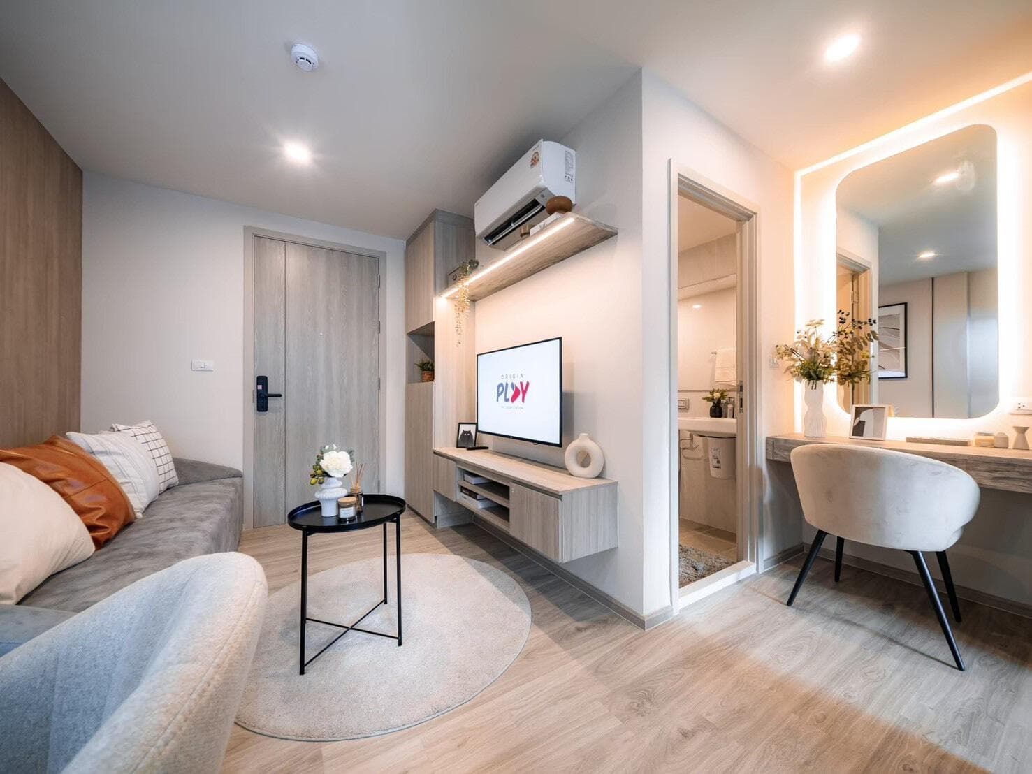 Show unit photograph of a 28 sqm Smart Kitchen unit showing the living area with wall-mounted television, the entrance door with digital lock and the lit vanity beside the bathroom.