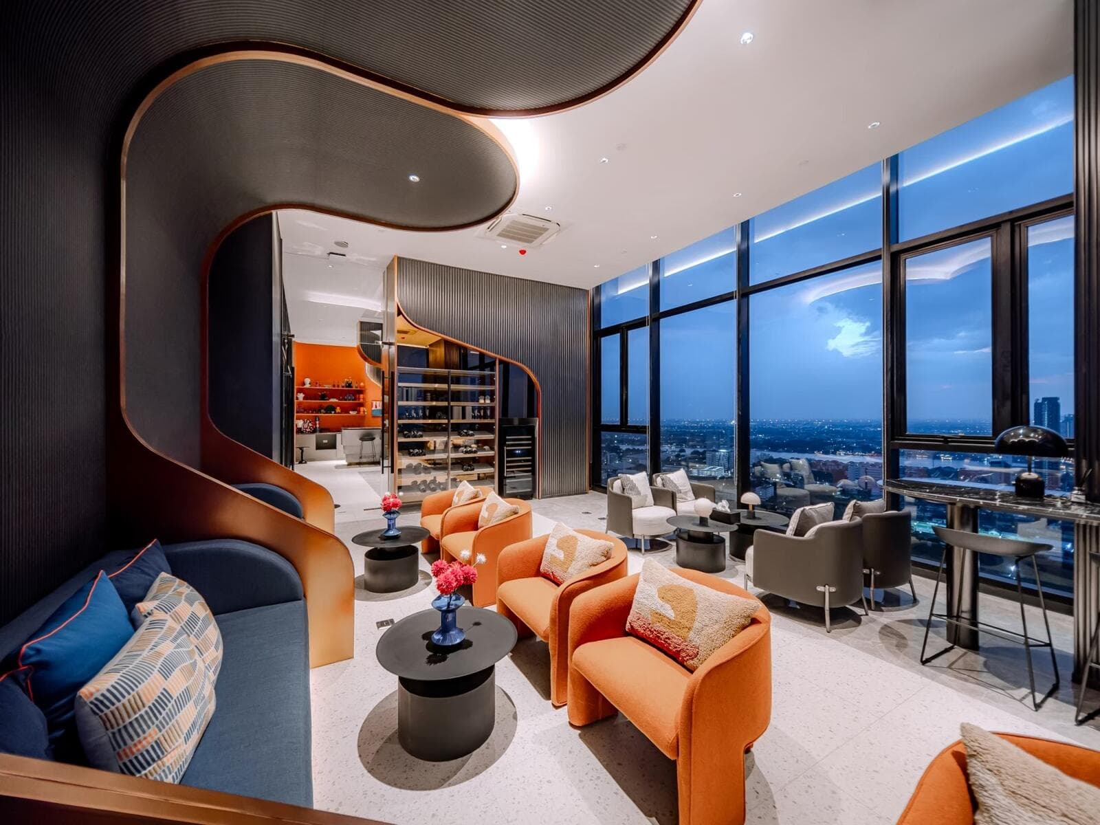 Photograph of the sky lounge at dusk with armchair seating, a pantry and display shelving, and floor-to-ceiling windows over Nonthaburi.