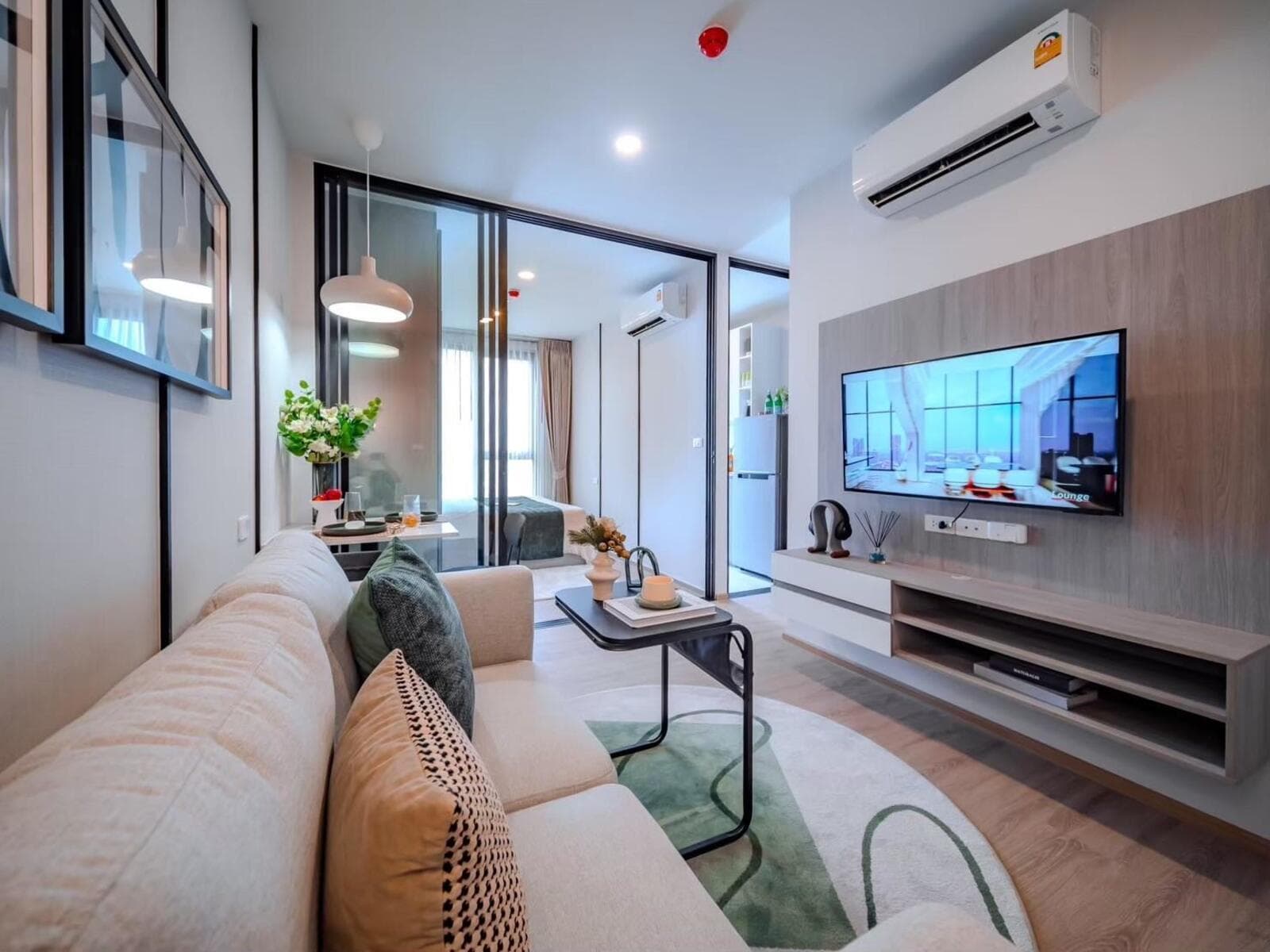 Show-unit living area of a 26 sqm one-bedroom layout, with a fabric sofa, wood-panelled television wall and a glass partition to the bedroom.