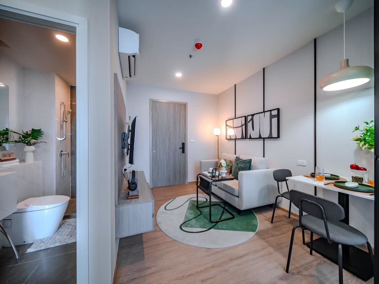 Living and dining area of a furnished show unit at Origin Plug & Play Nonthaburi Station, with a round green rug, a dining table and the bathroom door ajar