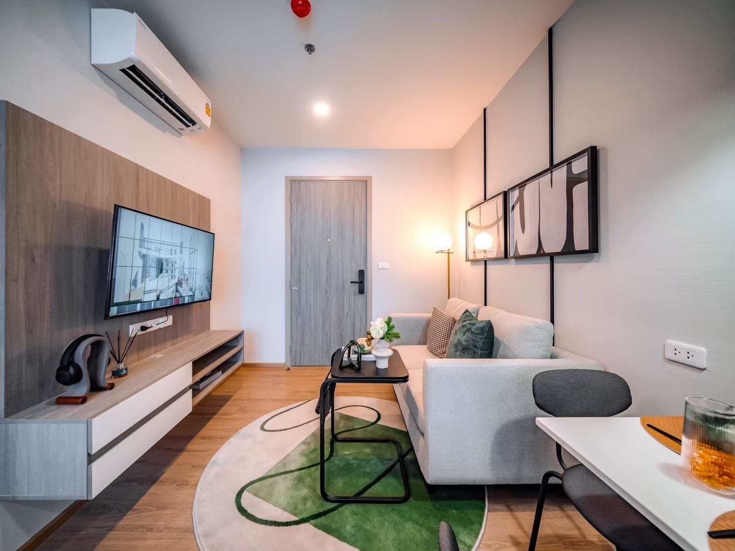 Living area of a furnished show unit at Origin Plug & Play Nonthaburi Station, with a wall-mounted television, a pale sofa and a timber panelled wall