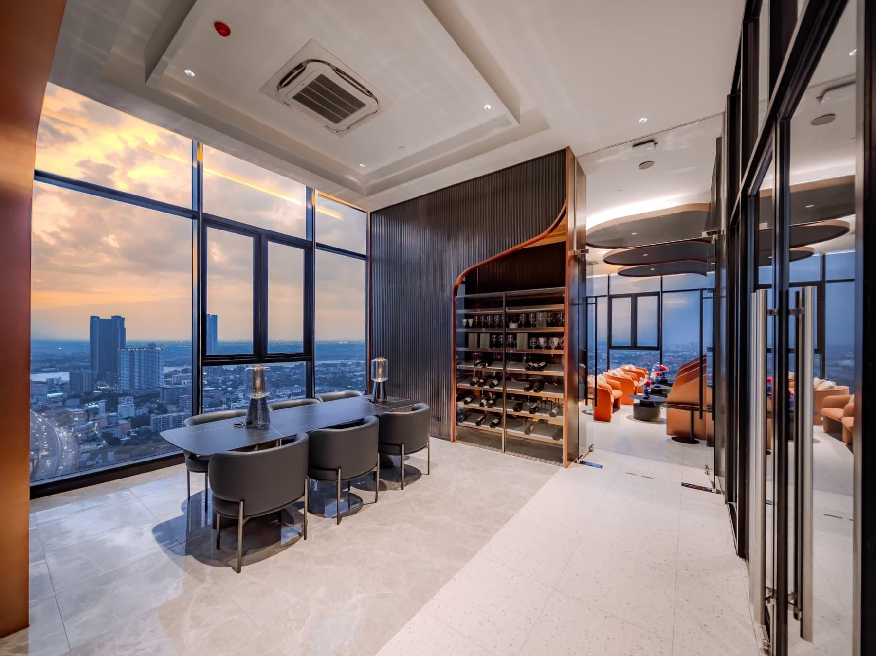 Sky lounge at Origin Plug & Play Nonthaburi Station, with a dining table, a wine display wall and full-height glazing over the city at dusk