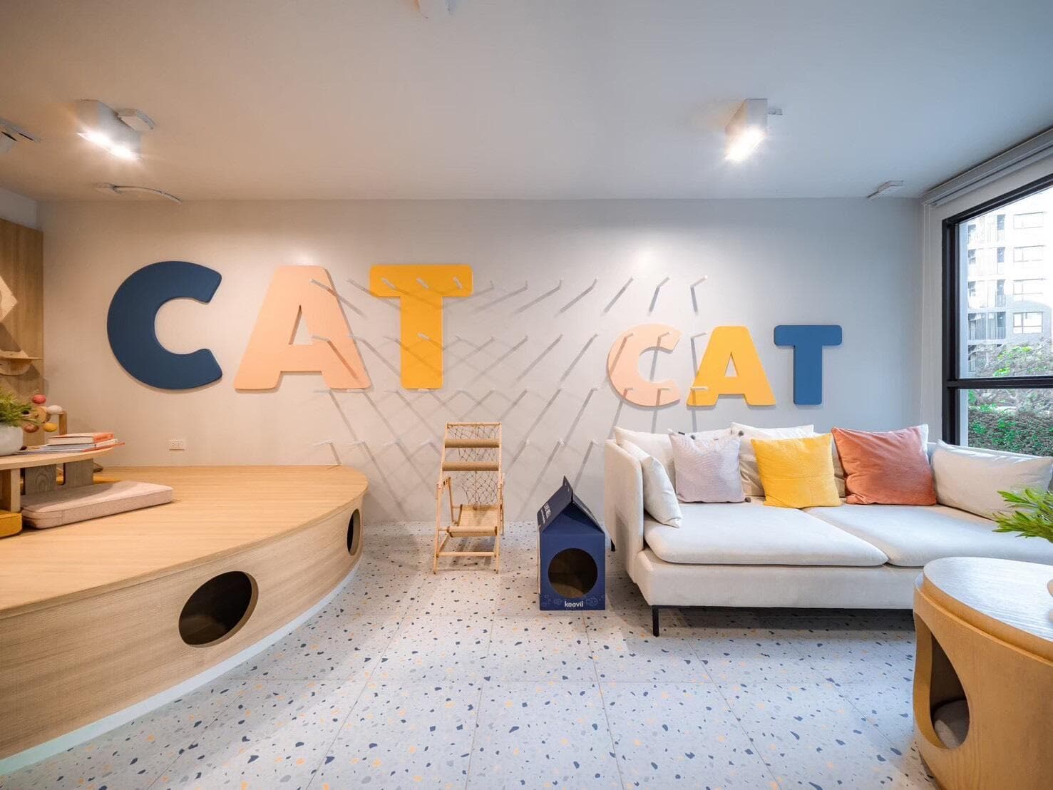 Photograph of the indoor cat lounge with a curved timber play platform, wall climbing steps, terrazzo flooring and a sofa beside the window.